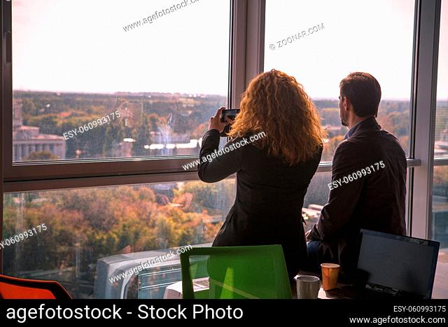 Business people resting after hard working day in office interior. Red haired lady making photos of city on mobile or smart phone