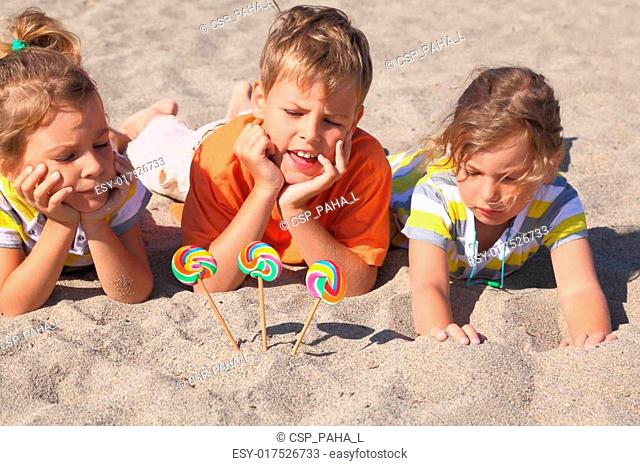 little boy and two girls lying on beach, chin on hands, lollipops stick in sand