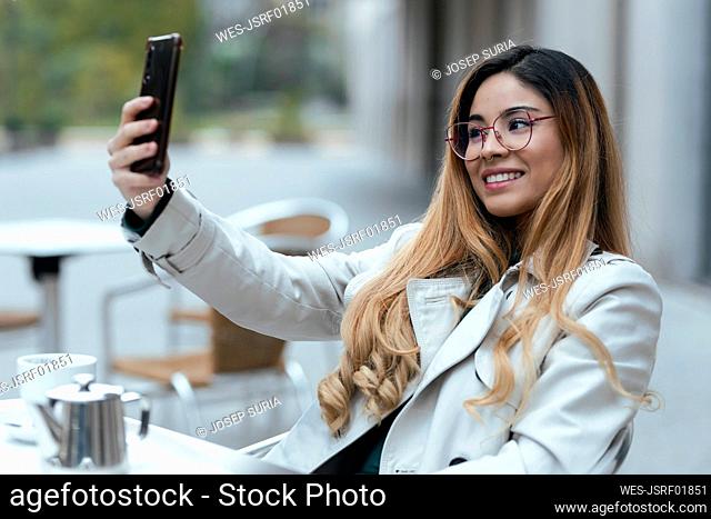 Smiling woman taking selfie through mobile phone sitting at sidewalk cafe