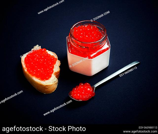 Still life with red caviar. Fresh caviar, sandwich, glass jar and spoon on black background