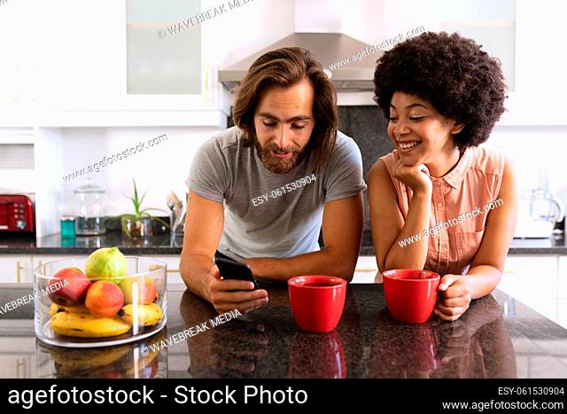 Happy diverse couple standing in kitchen drinking coffee and using smartphone