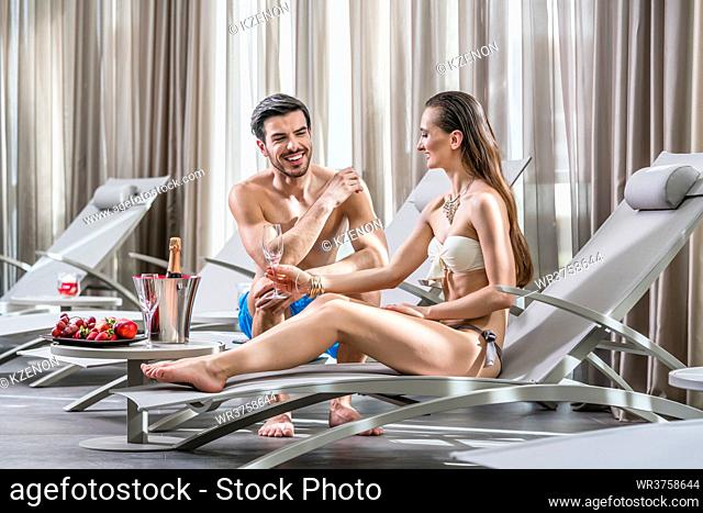 Romantic young man opening a bottle of champagne while relaxing with his partner at the swimming pool in a fancy hotel