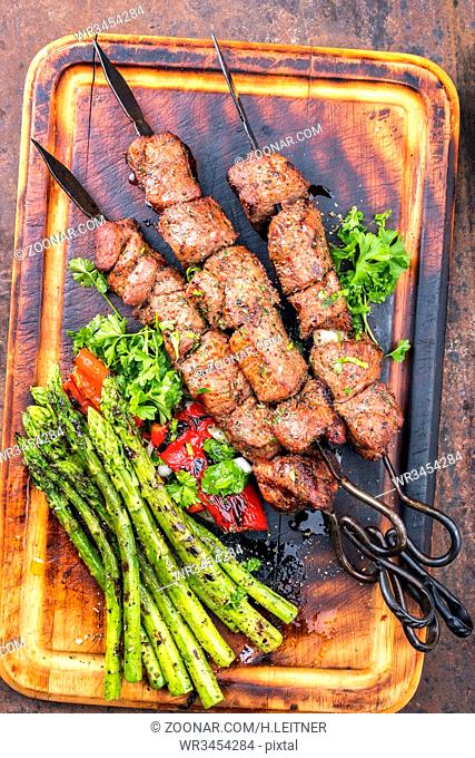 Traditional Russian shashlik on a barbecue skewer with green asparagus and paprika as top view on a burnt cutting board