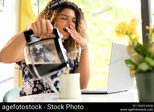 Tired woman pouring coffee by laptop