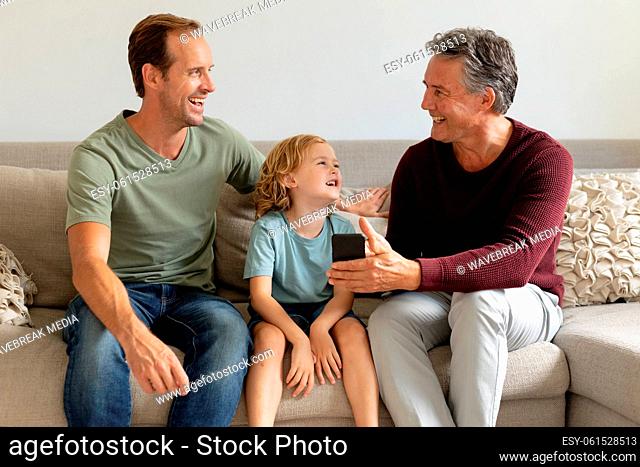 Senior caucasian grandfather on couch looking at smartphone with adult son and grandson laughing