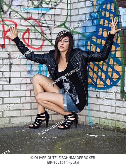 Young woman with dark hair wearing hot pants, a black leather jacket and high heels posing in front of a wall with graffiti