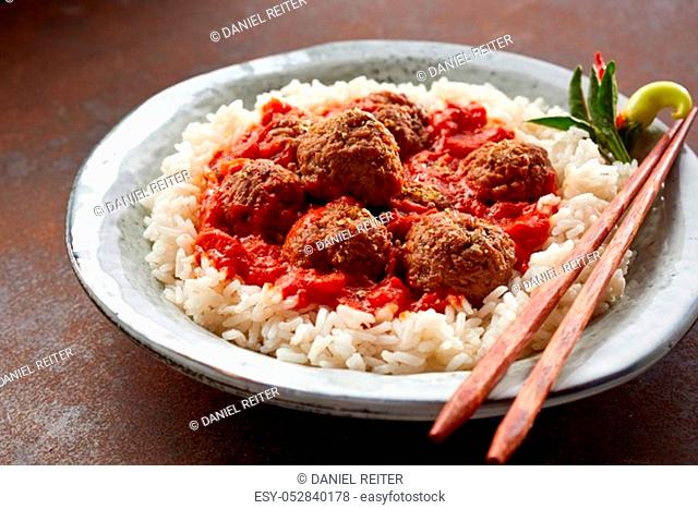 Angled close up on meatballs bowl containing thai rice and several meatballs in sauce on top of scratched red surface