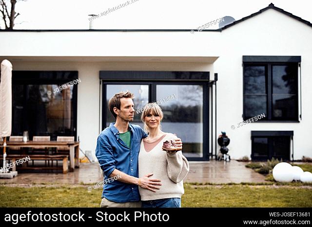 Man touching belly of pregnant woman holding baby booties in front of house
