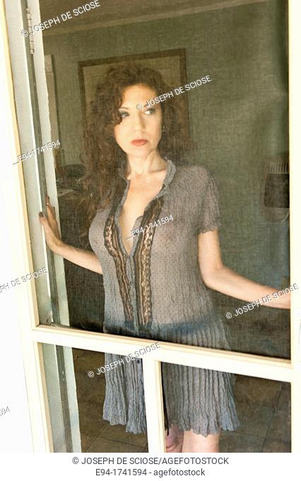 Portrait of a 42 year old woman wearing a sheer dress with long curly black hair looking away from the camera in a provocative pose standing behind a screen...