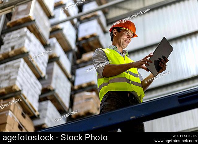 Smiling worker wearing hardhat using tablet PC by railing in warehouse