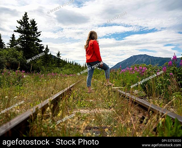 Woman walking down the railroad tracks in the Yukon with Fireweed (Chamaenerion angustifolium) blooming all around; Carcross, Yukon, Canada