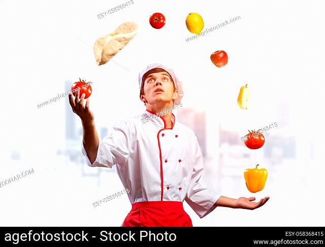 Young male chef juggles with food ingredients. Handsome chef in white hat and red apron in light kitchen interior. Professional cooking classes advertising