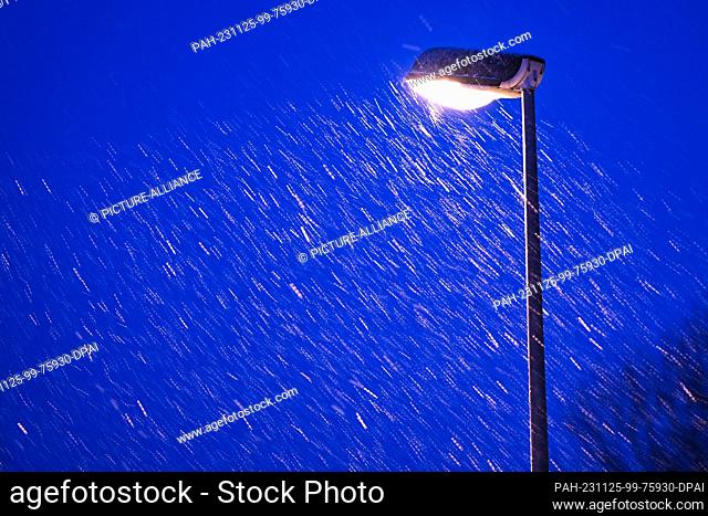 25 November 2023, Baden-Württemberg, Isny im Allgäu: Snowflakes blow around a street lamp at the blue hour. It started snowing in the Allgäu shortly after...