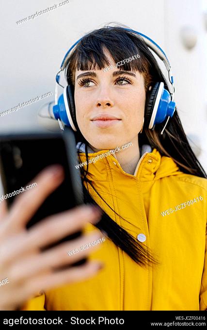 Young woman with headphones and mobile phone looking away while standing outdoors