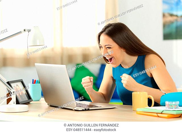 Single excited student on line with a laptop sitting in a desk in her room in a house interior