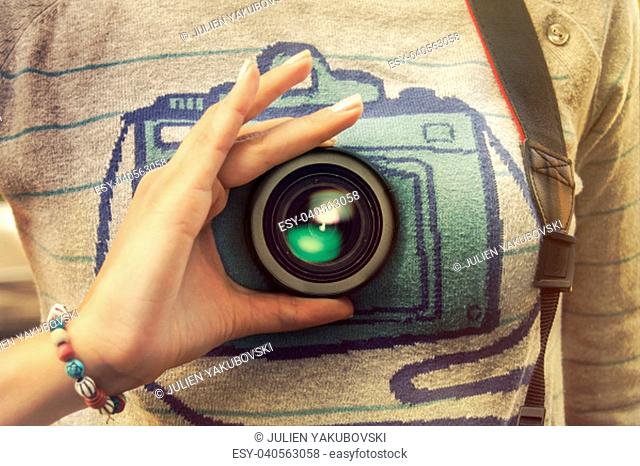 A young woman hand holding camera lense infront of her brest.She wears a sweater with a camera print