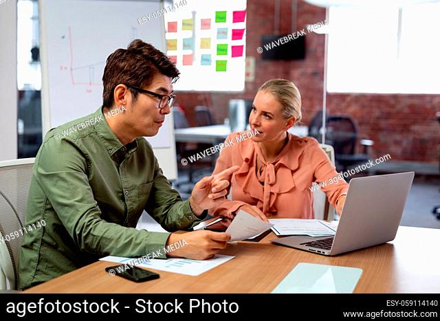 Diverse male and female colleague sitting at table with laptop and paperwork discussing