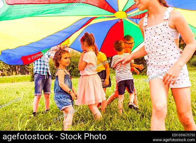 Portrait of adorable little girl standing unsure, trying to join active game of older kids. Children playing tag game under large rainbow canopy