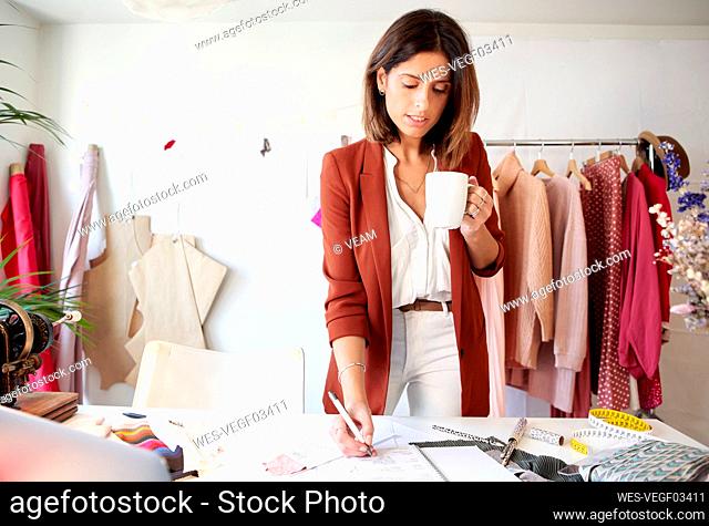 Beautiful fashion designer writing in diary while holding coffee cup against clothes rack at studio