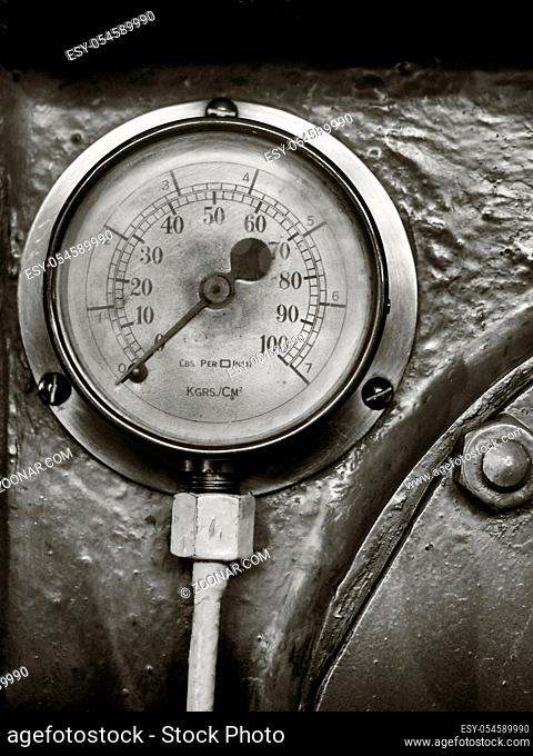 sepia monochrome image of an old shiny brass round pressure gauge with a round dial marked in numbers on a metal panel