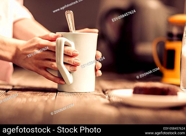 Female hands holding a white cup of tea standing close to wooden desk. Close up view of mug with teaspoon inside