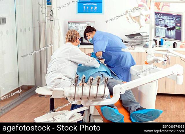 Senior dentist woman performing surgery putting implant into sick man mouth in dental clinic. Dentistry doctor with protection mask treating teeth of patient...