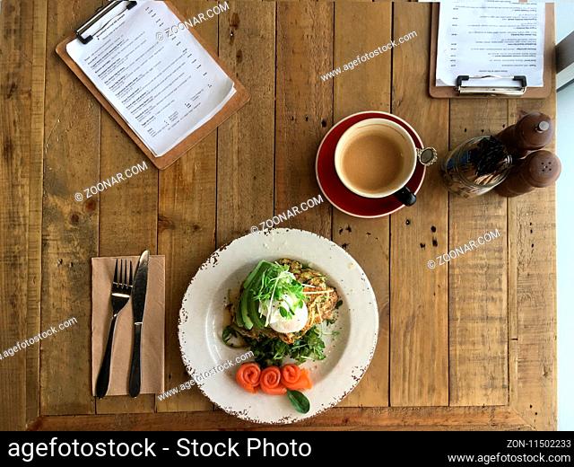 Top view looking down on a rustic table and a plate of zucchini and sweet potato fritters, poached eggs, avocado and rolled rosette salmon on the side with a...