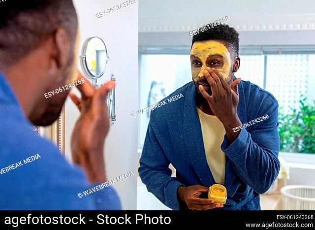 African american young man looking at mirror and applying facial mask in bathroom