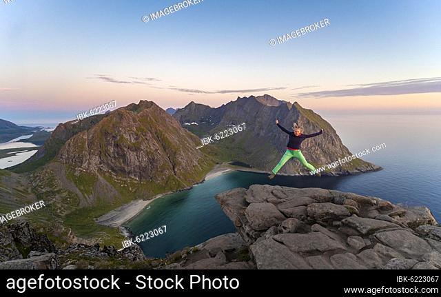 Young woman making a jump in the air, on top of Ryten, sea, Kvalvika beach and mountains, Fredvang, Lofoten, Nordland, Norway, Europe