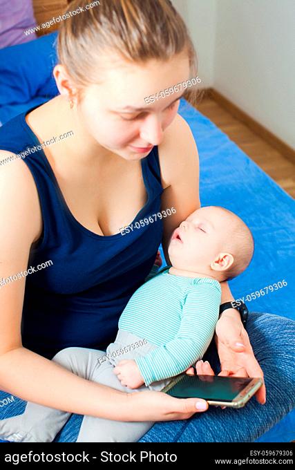 Lovely woman checking messages on her smartphone while small cute baby is sleeping on her knees. Mother using new application on her phone