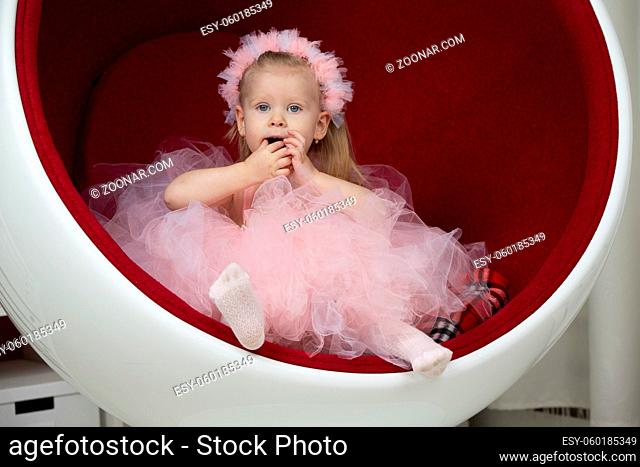 Two-year-old child. A smart little girl in a pink fluffy dress sits on a red chair