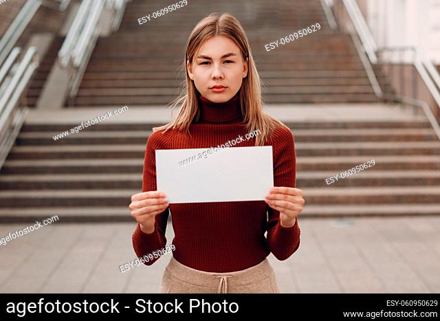 Young woman holds white paper in hand stair street background. Two women talking at table in street cafe. White blank template sheet with empty space