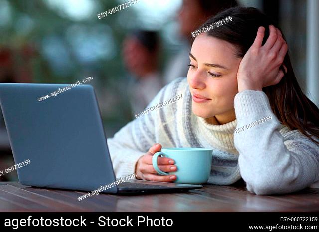 Happy woman holding coffee mug checking laptop content in winter in a terrace