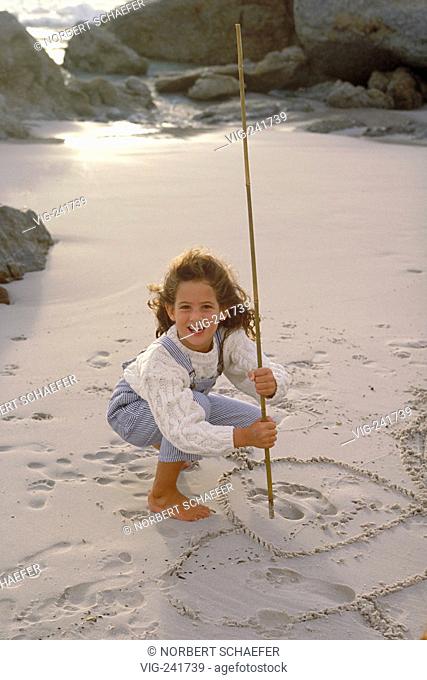 beach-scene, bare feeted 8-year-old girl wearing striped trousers with braces and a white pullover draws with a stick into the wet sand  - GERMANY, 26/06/2004