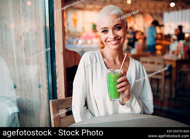 Portrait of young smiling millenial european short haired woman with green smoothie at cafe. Beautiful happy blonde girl indoor