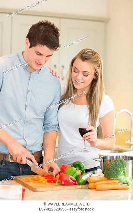 Man cutting peppers with woman drinking wine in kitchen