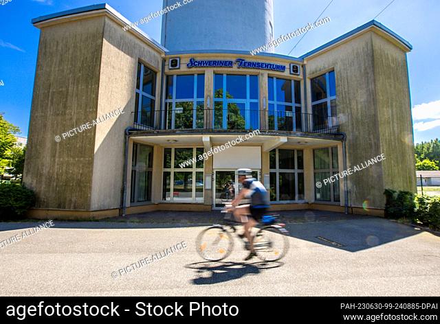 PRODUCTION - 30 May 2023, Mecklenburg-Western Pomerania, Schwerin: A cyclist rides past the entrance building to the TV tower