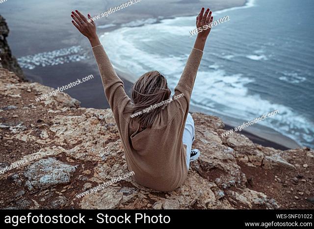 Carefree woman with arms raised sitting on mountain against Famara Beach, Lanzarote, Spain