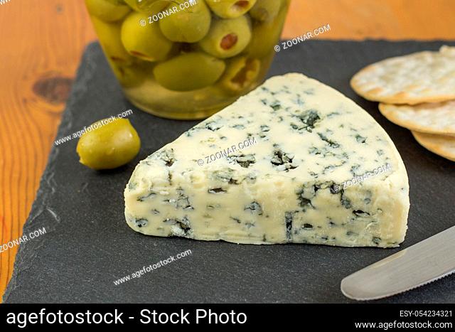 Blue cheese wedge with defocused jar of green olives and crackers in background
