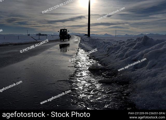 09 December 2023, Bavaria, Ruderatshofen: Ice thaws on a road in a wintry landscape against a panorama of the Alps in the sunshine