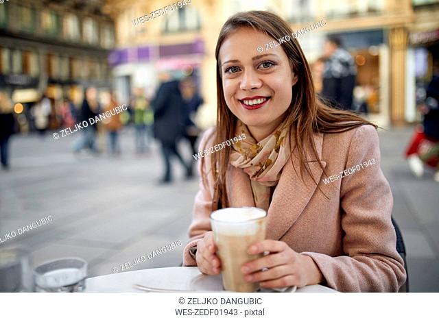 Austria, Vienna, portrait of smiling young woman drinking coffee at pavement cafe