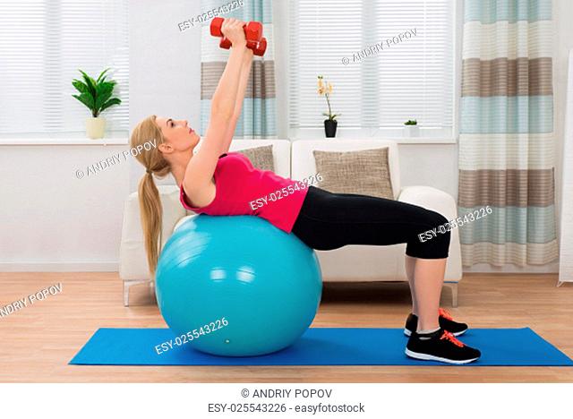 Young Woman Holding Dumbbell While Exercising On Fitness Ball In Living Room
