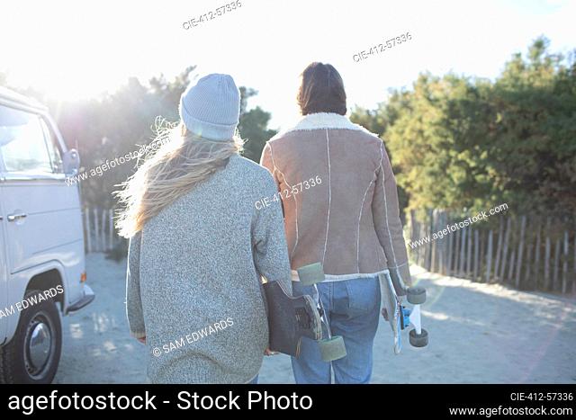 Young women friends with skateboards walking on sunny beach path