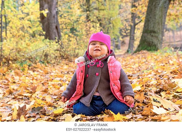 Happy baby girl in pink warm coat and pink hat makes grimace sitting on fallen leaves in beautiful autumn park on fall day