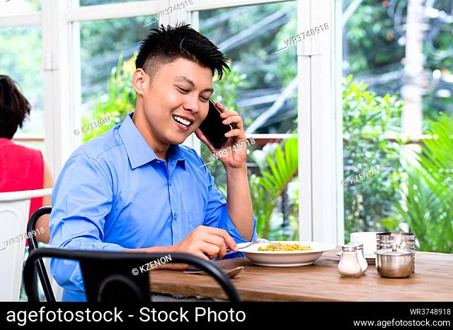 Young Asian man chatting on his mobile phone as he sits at a table in a restaurant with a plate of food in front of him smiling as he listens to the...