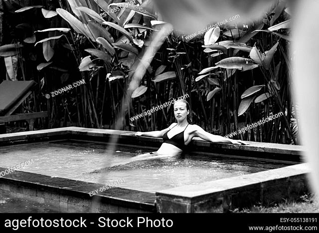 Sensual young woman relaxing in outdoor spa infinity swimming pool surrounded with lush tropical greenery of Ubud, Bali. Wellness