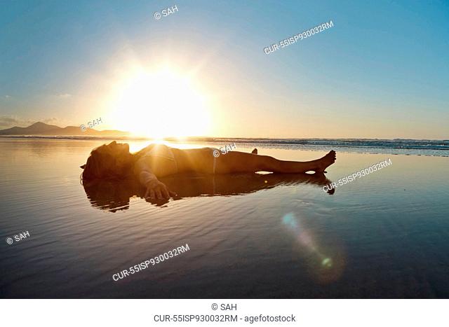 Mid adult woman lying in shallow water on beach at sunset
