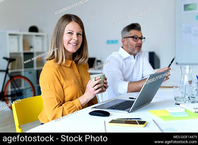 Smiling businesswoman holding cup sitting at desk with colleague at office