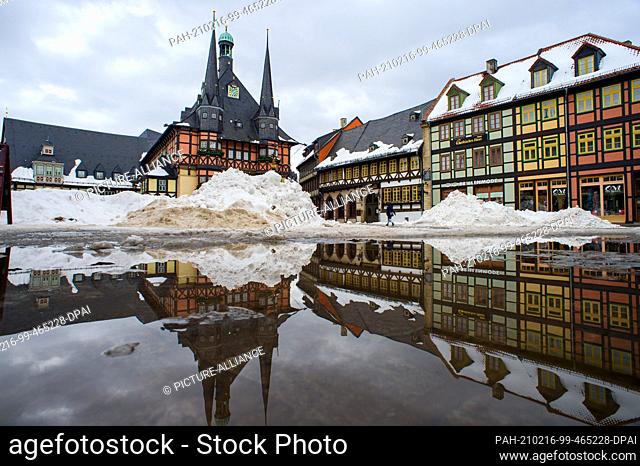 16 February 2021, Saxony-Anhalt, Wernigerode: Mountains of snow pile up on the market square, while the town hall and the adjacent houses are reflected in the...