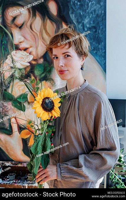 Smiling female artist holding sunflower while standing against artwork